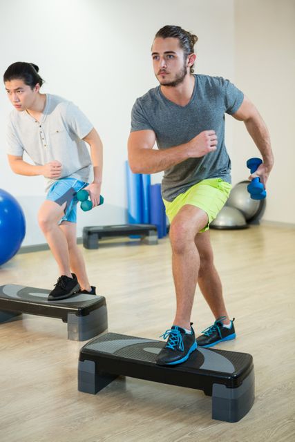 Two men are engaging in a step aerobics workout using dumbbells in a fitness studio. They are focused and determined, performing the exercise on steppers. This image is ideal for promoting fitness classes, gym memberships, workout routines, and healthy lifestyle campaigns.