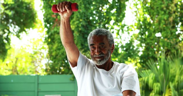 Elderly man performing workout routine with dumbbell in lush garden background. Shows dedication to active and healthy lifestyle amidst natural surroundings. Ideal for articles on senior fitness, outdoor exercise routines, and promoting health among older generations.