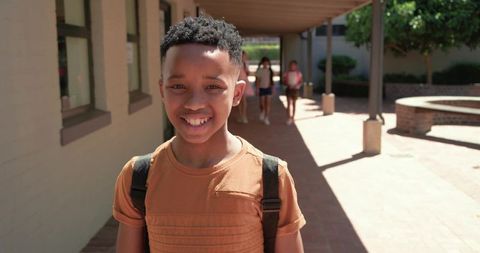Smiling black teenage student with backpack walking sunlit campus corridor with classmates