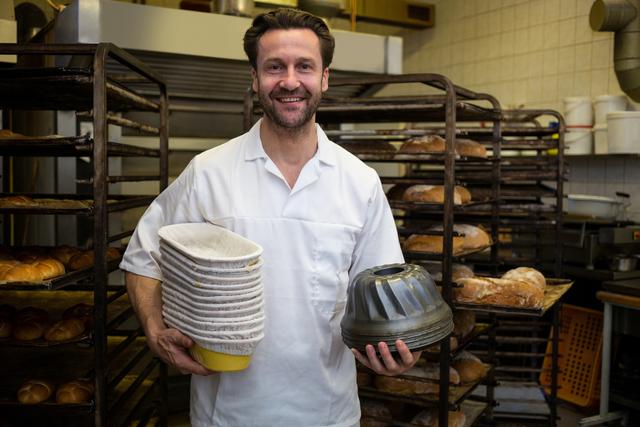 Smiling baker holding mould and stack of boxes in bakery kitchen. Ideal for use in articles about baking, culinary arts, professional chefs, and the food industry. Suitable for illustrating concepts of artisanal baking, workplace happiness, and professional kitchen environments.