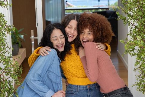 Three friends hugging and laughing in cozy doorway with indoor plants and casual style
