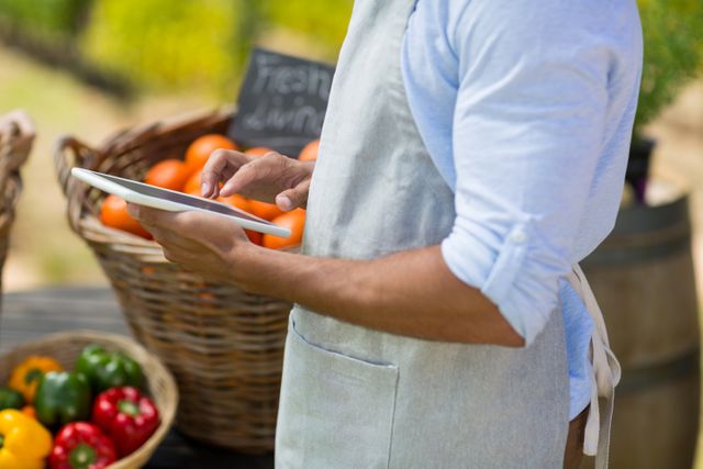 Farmer wearing apron using digital tablet at outdoor farm market with baskets of fresh vegetables. Ideal for illustrating modern farming, technology in agriculture, small business operations, and local organic produce marketing.