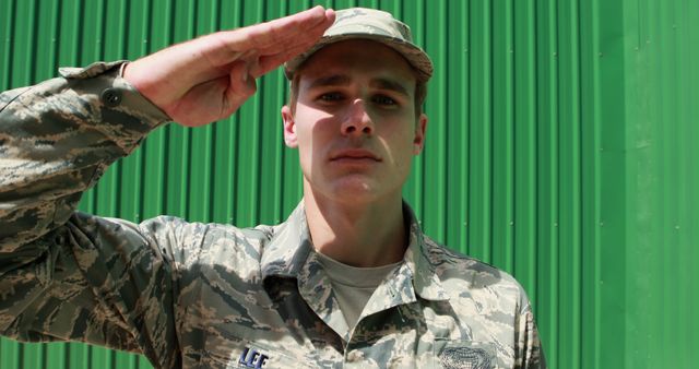 A young male soldier in camouflage fatigues giving a salute in front of a green wall. Can be used for military recruitment campaigns, patriotic events, army advertisements, or information on national service. Ideal for educational purposes highlighting armed forces and their role in national security.