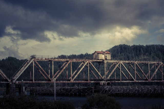 An abandoned railroad bridge stretches over a river under a cloudy sky. The structure is adorned with weathered metal and rust, enhancing its aged and rustic appearance. This image can be used effectively for themes related to dereliction, history, industrial heritage, or nature reclaiming man's constructions. It is also suitable for backgrounds in design projects or storytelling needing a moody or vintage-industrial setting.