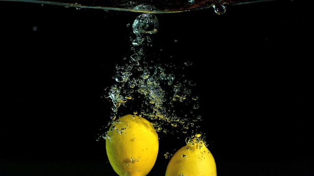 Two vibrant yellow lemons creating splash in clear water with bubbles rising against black background. Suitable for food and beverage promotions, health and wellness articles, refreshing summer themes, fruits marketing, and creative advertising campaigns.