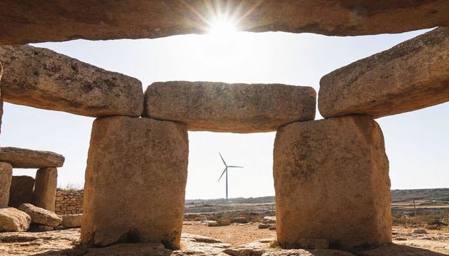 Megalithic trilithon framing wind turbine with sunburst on rural limestone plateau