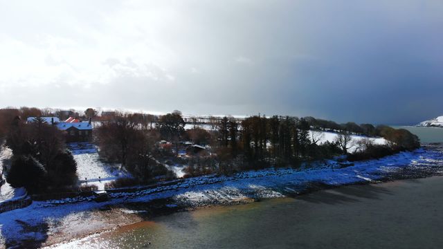 Aerial view of a picturesque winter scene with a serene river and surrounding snow-covered countryside. Overcast sky adds dramatic effect. Ideal for themes of tranquility, nature's beauty, winter travel, and serene retreats.