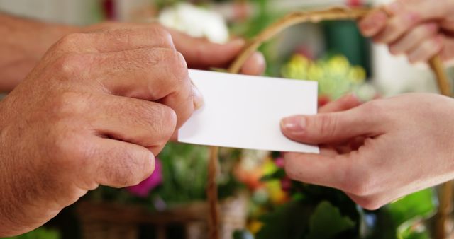 Close-up view of hands exchanging a white business card over a vibrant floral arrangement. Useful for representing networking, business communication, professional interactions, and contact information exchange in various marketing and business contexts.