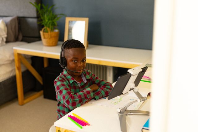 Young African American boy wearing headphones, using a tablet at home. He is smiling and appears to be engaged in an online learning activity. The setting includes a desk with colorful markers and a plant in the background, suggesting a comfortable and organized study space. Ideal for illustrating concepts of remote learning, online education, childhood technology use, and happy domestic life.
