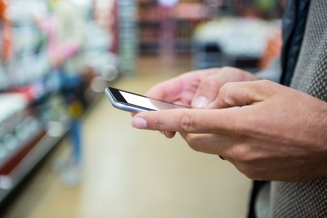 Man's hands holding and using a mobile phone in a supermarket. Ideal for illustrating modern shopping habits, technology in retail, and consumer behavior. Useful for articles on mobile technology, retail trends, and convenience in daily life.