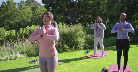 Diverse group practicing outdoor yoga on mats in sunny park meditating with hands in prayer