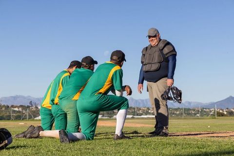 Kneeling baseball players listening to smiling catcher in protective gear on sunlit field