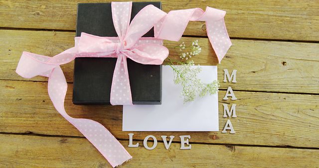A gift box adorned with a pink ribbon and a note with the words MAMA LOVE rests on a wooden surface, symbolizing a thoughtful present for Mother's Day. The scene captures a heartfelt gesture of appreciation and affection towards a mother.