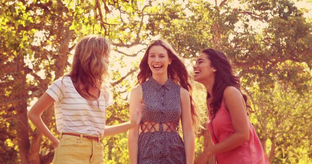 Three young women are laughing together outdoors in a sunlit environment. They wear casual summer clothing and appear to be enjoying each other's company. This image is perfect for promoting outdoor activities, friendship, and positive emotions. It can be used in advertisements for lifestyle brands, social media campaigns, and content related to summer activities and youthfulness.