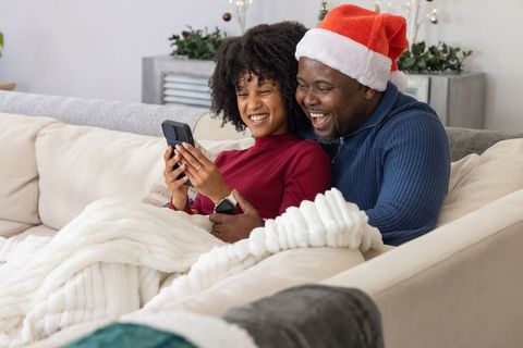 African american father and teen daughter sharing holiday laughter on couch with phone
