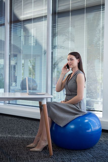 Female executive sitting on exercise ball while talking on mobile phone at desk