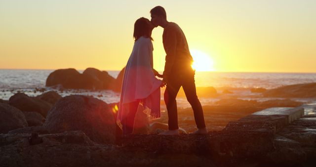 A young Caucasian couple shares a romantic moment at sunset on a rocky beach, with copy space. Their silhouettes against the vibrant sky create a serene and intimate atmosphere.