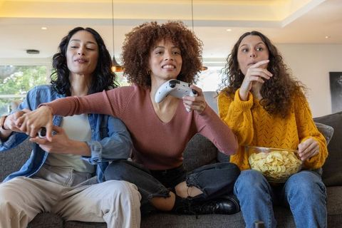 Diverse women playing console game on couch laughing and sharing snacks