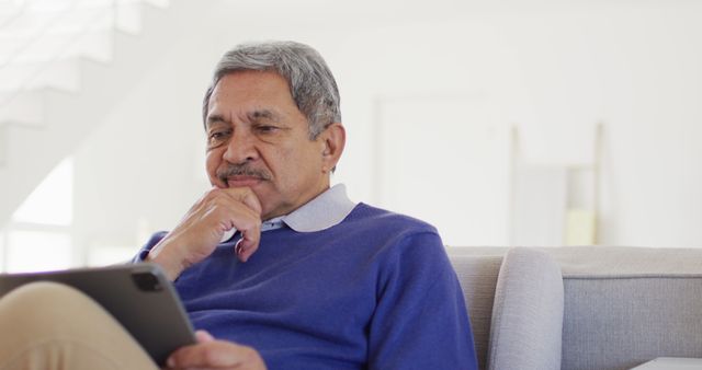 Elderly man is using tablet while sitting on couch in a modern, bright living room. Image can be used for tech adoption by seniors, leisure activities, or digital literacy campaigns.