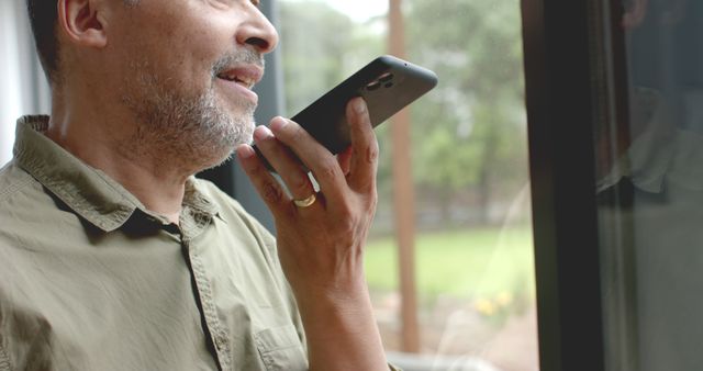 Mature man using voice assistant on smartphone near window, exemplifying use of modern technology in daily life. Useful for themes of technology, communication, and senior lifestyle. Can be used in articles or advertisements related to innovations in mobile communication, age-friendly technology, or home ambiance.