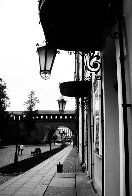 Classic black and white photo capturing a quiet street adorned with ornate lanterns and an ancient archway in the background. Ideal for use in historical-themed projects, vintage decor, articles about architecture or urban landscapes, and travel blogs highlighting historic cities.