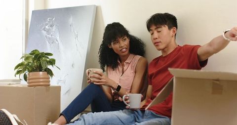 Young diverse couple relaxing with mugs while unpacking moving boxes in cozy apartment
