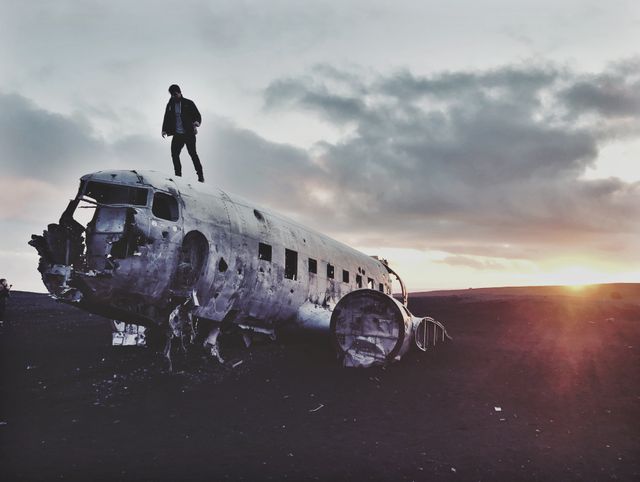 This visual captures an individual standing on the wreck of an old airplane in a deserted, desolate area during sunset. The dramatic clouds and horizon add an artistic and adventurous feel to the scene. Perfect for themes related to adventure, exploration, solitude, travel, and outdoor landscape photography.