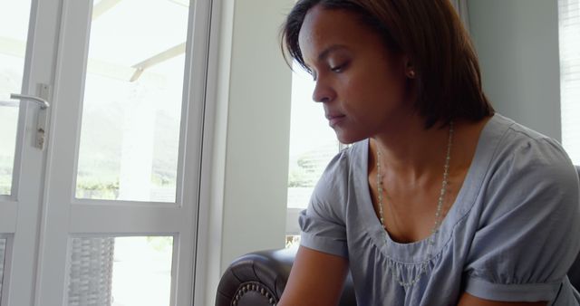 A woman sits on a sofa near a window with natural light shining in, appearing deep in thought. Suitable for topics such as relaxation, mental health, personal reflection, moments of solitude, or lifestyle content.