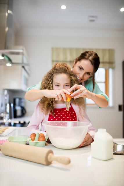 Mother and daughter baking together in a modern kitchen, with the mother assisting her daughter in breaking eggs into a mixing bowl. The kitchen counter is filled with baking ingredients and utensils. Perfect for themes related to family bonding, home baking, cooking education, and parent-child activities.