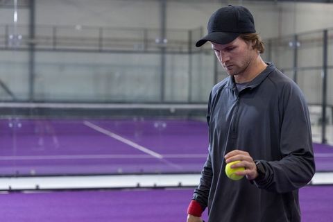 Young man concentrating while holding padel ball on purple indoor court with glass panels