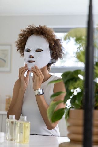 African american woman wearing led face mask at vanity applying skincare routine for glow