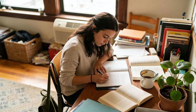 College student studying at sunlit wooden desk with open books and coffee mug