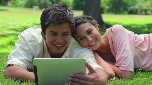 A couple lying on grass in the countryside, happily watching a tablet. Ideal for lifestyle, technology in nature, outdoor relaxation, digital entertainment, and couple bonding themes.