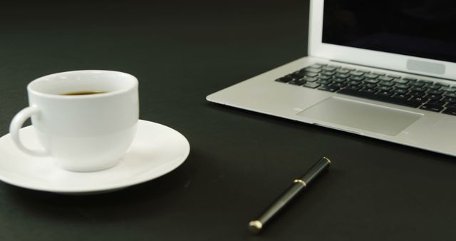 A white coffee cup sits next to an open laptop on a dark surface, with copy space. This setup suggests a professional or academic environment, involving work or study.