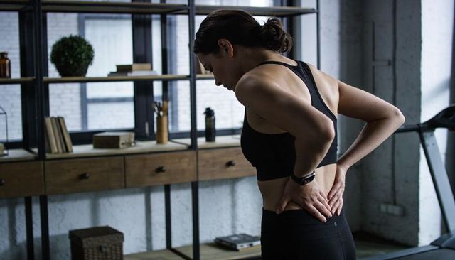 Woman holding lower back and leaning in home gym near treadmill and minimalist shelving