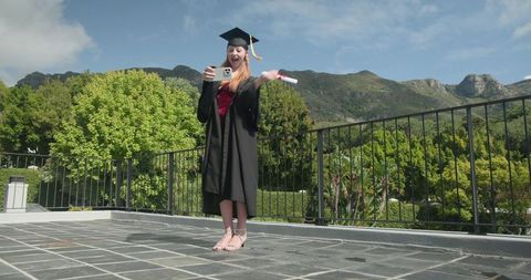 Graduate celebrating on terrace holding diploma and smartphone during virtual ceremony