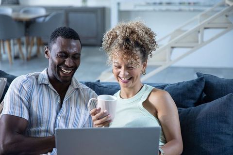Laughing black man and biracial woman relaxing on sofa with mug, watching laptop together