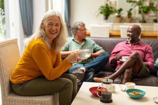 Senior woman smiling while holding coffee cup, sitting with multiracial male friends in living room. Ideal for depicting social gatherings, leisure time, and senior lifestyle. Useful for articles on friendship, diversity, and home life.