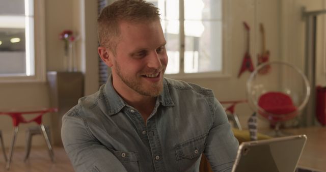 Young man in casual denim shirt smiling while using a digital tablet in a modern office. Ideal for business, remote work, technology, and teamwork concepts. Office setting with contemporary furniture and daylight provides a casual yet productive atmosphere.