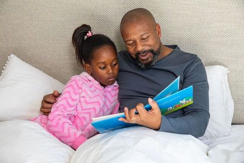 African american father reading bedtime story with daughter cuddling in pink pajamas