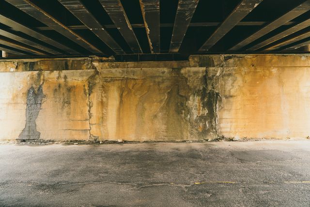 Weathered concrete wall under overpass displaying aging and textured surface. Ideal for urban, industrial, and infrastructure themes, or as a backdrop for grunge and alternative art projects.