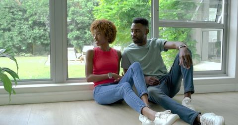 Diverse couple relaxing on sunlit wood floor by floor-to-ceiling window with backyard view