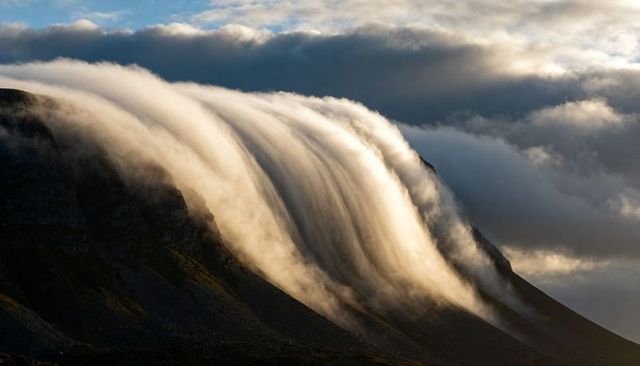 Cascading clouds spilling over jagged mountain ridge in golden light at sunrise