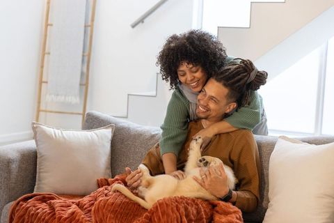 Multiracial couple cuddling cream dog on cozy sofa, sharing warm intimate home moment