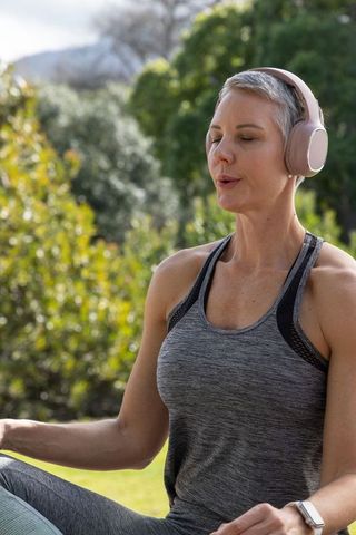 Middle-aged woman meditating outdoors wearing pink headphones and smartwatch for wellness