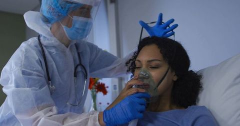 Nurse wearing full ppe administering oxygen therapy and positioning mask for patient in hospital bed