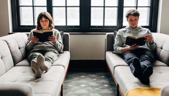 Couple reclining on sofas reading by window during cozy at-home afternoon