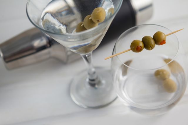 High angle view of a vodka martini garnished with olives, accompanied by a cocktail shaker on a wooden table. Ideal for use in bar and restaurant promotions, cocktail recipes, and lifestyle blogs focusing on drinks and mixology.