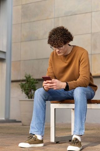 Young man sitting on bench using smartphone wearing glasses casual sweater urban courtyard