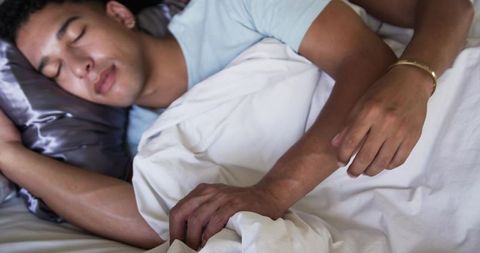 Sleeping young man resting under white duvet with satin pillow and gold bracelet details